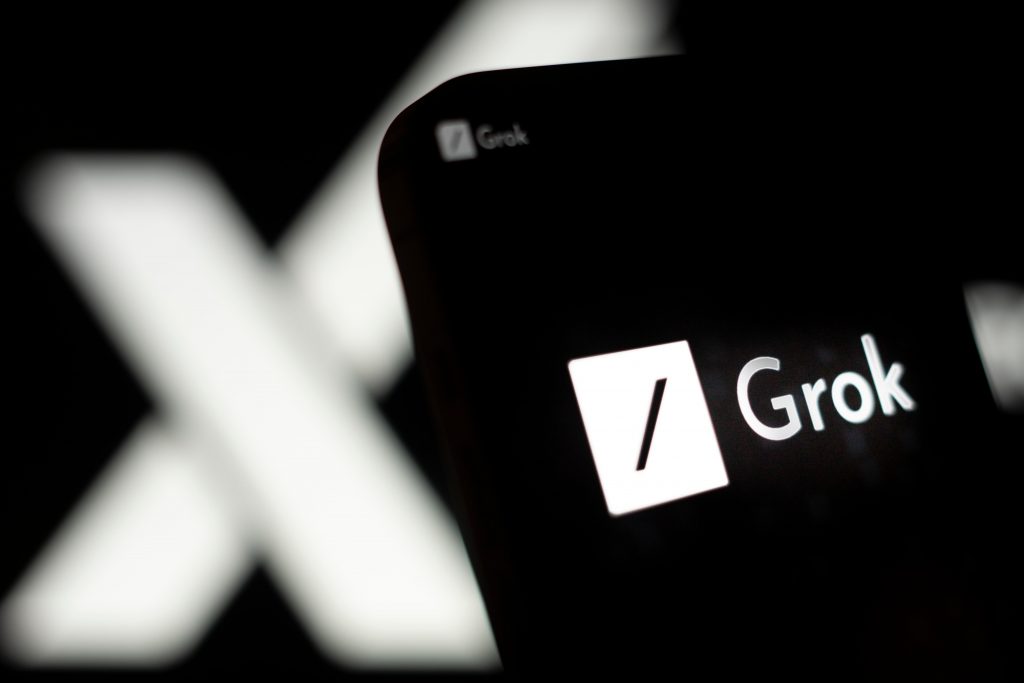 Pantalla de un dispositivo móvil que muestra el logo de 'Grok' frente a una letra 'X' difuminada en el fondo.